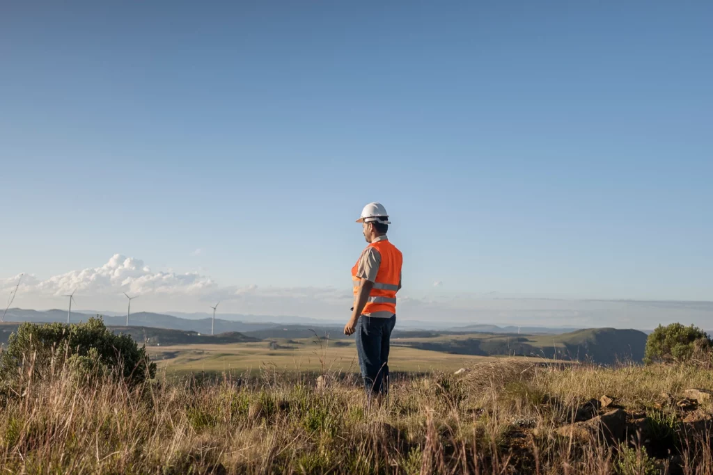 A worker in a safety vest and hard hat stands on a grassy hillside overlooking a wind farm and open landscape under a clear blue sky.