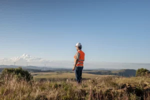 A worker in a safety vest and hard hat stands on a grassy hillside overlooking a wind farm and open landscape under a clear blue sky.